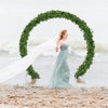 A woman in a flowing gown stands within an archway made of green ivy garlands on a beach.