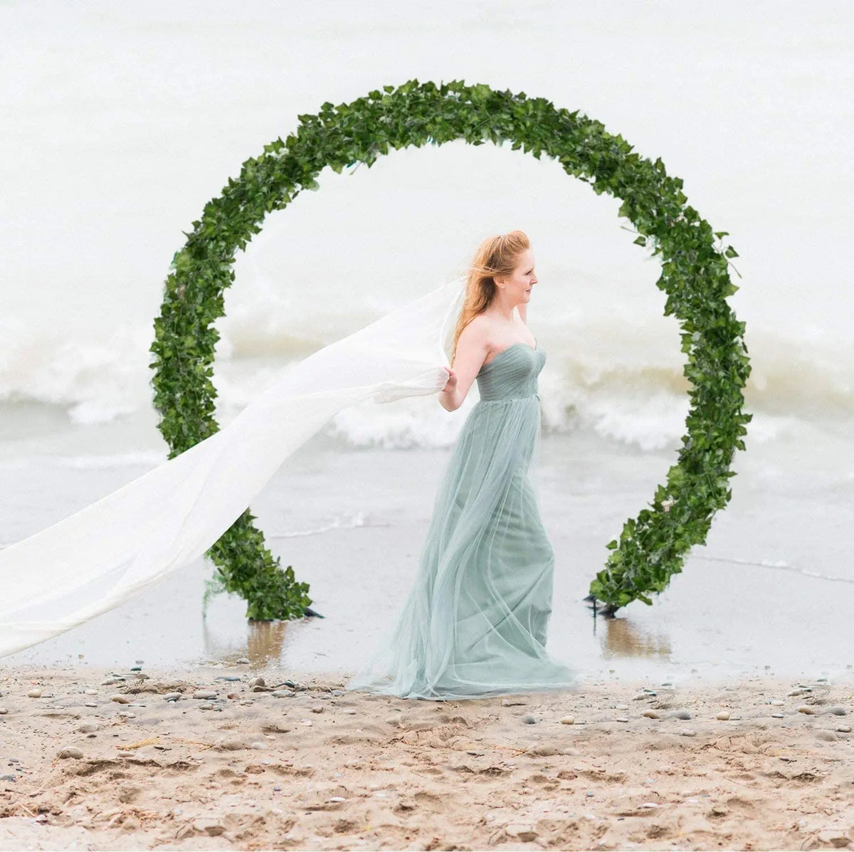 A woman in a flowing gown stands within an archway made of green ivy garlands on a beach.
