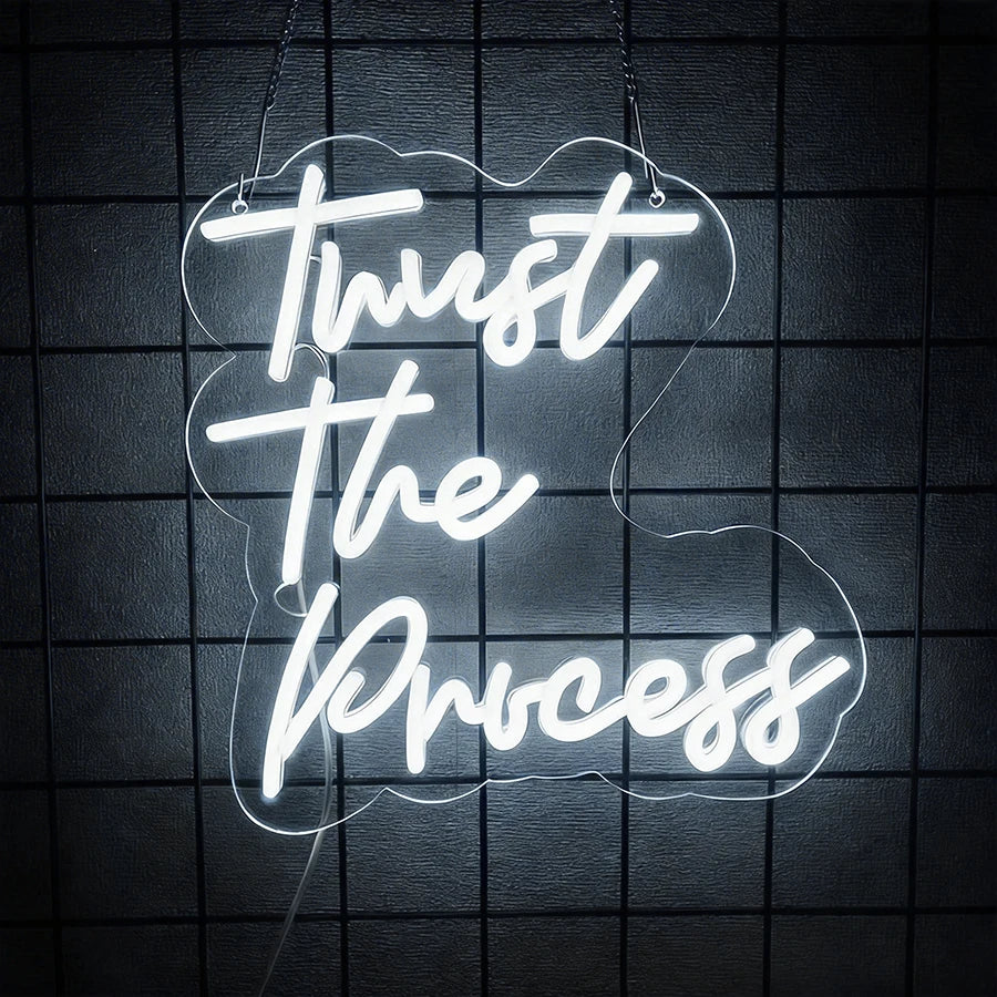 White neon sign on a dark surface displaying the text Trust The Process in a cloud shape.
