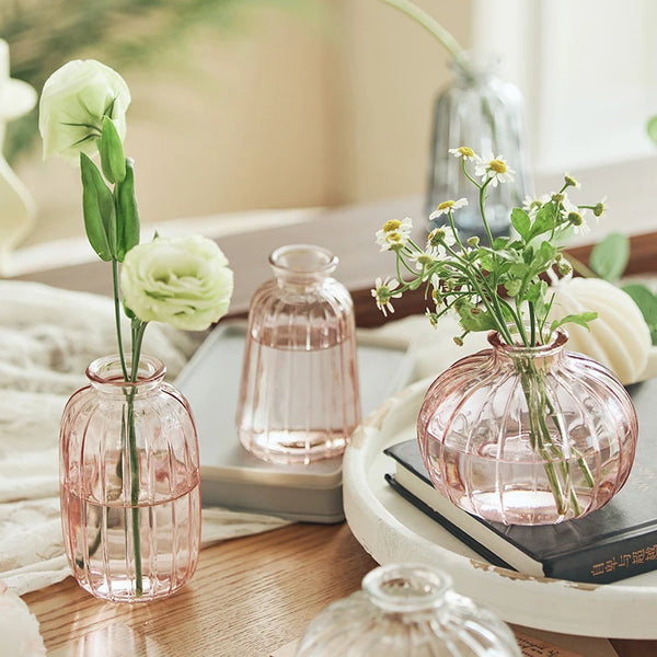 A pink textured glass vase filled with green flowers sits on a wooden table next to other glass items.