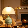 A retro farmhouse ceramic table lamp with a green crackle glaze sits on a wooden table in a warmly lit living room.