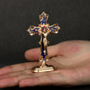 A gold metal crucifix with blue enamel details and a self-adhesive base is held in a person's hand.