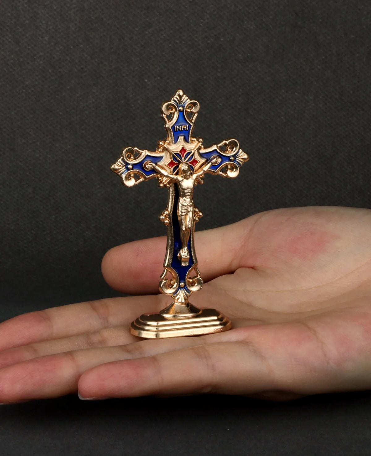 A gold metal crucifix with blue enamel details and a self-adhesive base is held in a person's hand.