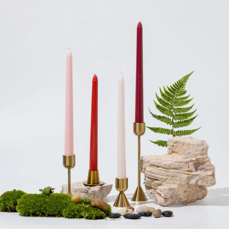 A collection of gradient color long taper candles in pink and red set against a natural stone and green moss background.