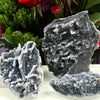 A cluster of dark gray natural zinc crystals displayed against a blurred background of green and red foliage.