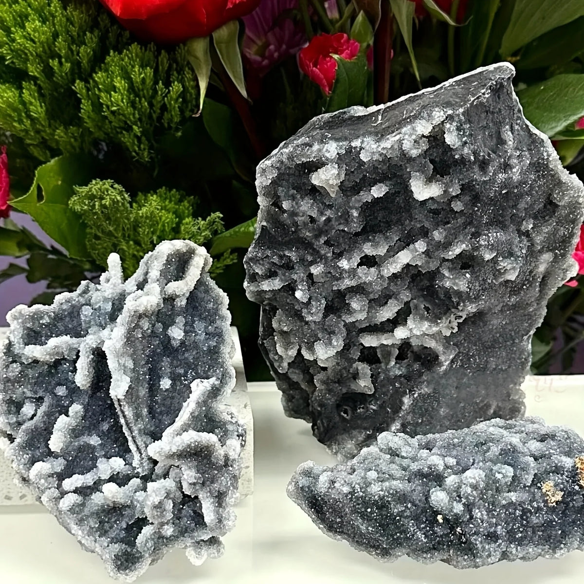 A cluster of dark gray natural zinc crystals displayed against a blurred background of green and red foliage.