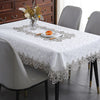 A white tablecloth with silver embroidered lace trim is set on a dining table surrounded by modern gray chairs.