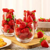 A glass bowl filled with bright red artificial strawberries on a wooden cutting board.