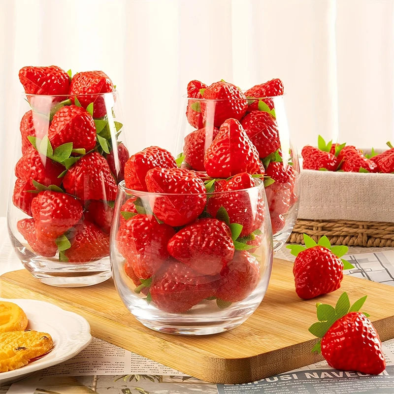 A glass bowl filled with bright red artificial strawberries on a wooden cutting board.