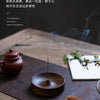 A dark wood incense stick burner with a thin metal stick is placed on a slate surface next to a small notebook and greenery.