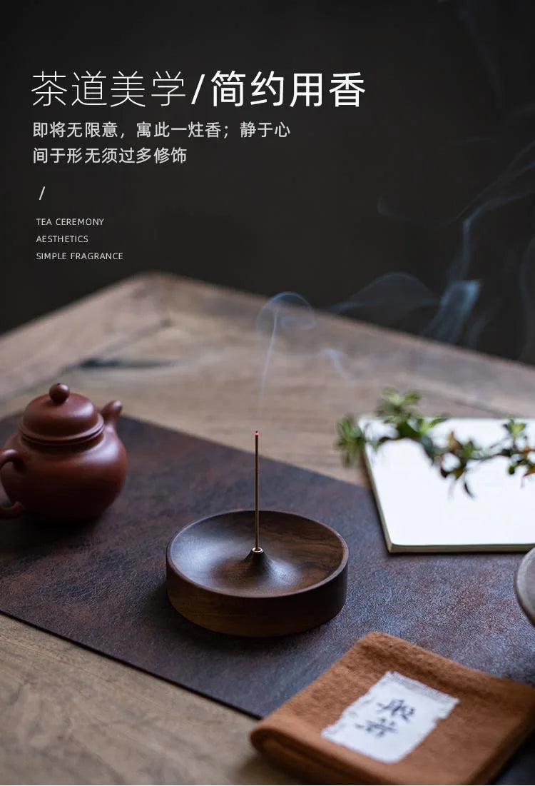 A dark wood incense stick burner with a thin metal stick is placed on a slate surface next to a small notebook and greenery.