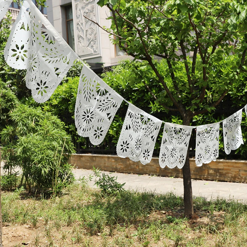 Eine Kette weißer, handgeschnittener Papel-Picado-Banner ist zwischen grünem Laub im Freien gespannt.