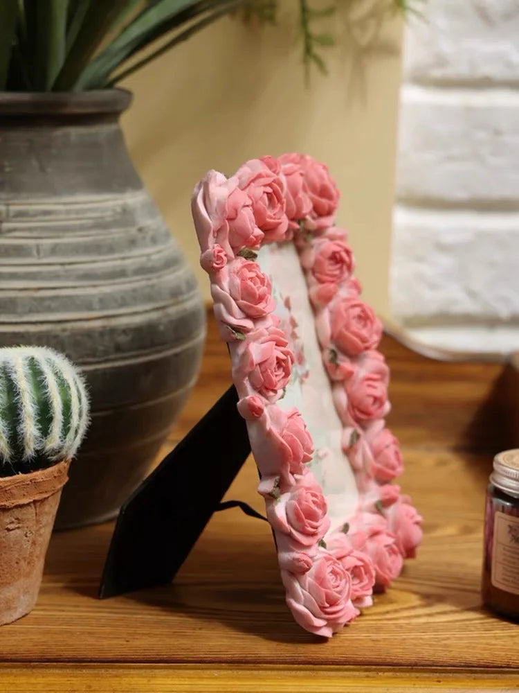 A pink rose resin picture frame is displayed on a wooden surface next to a potted plant and a gray vase.