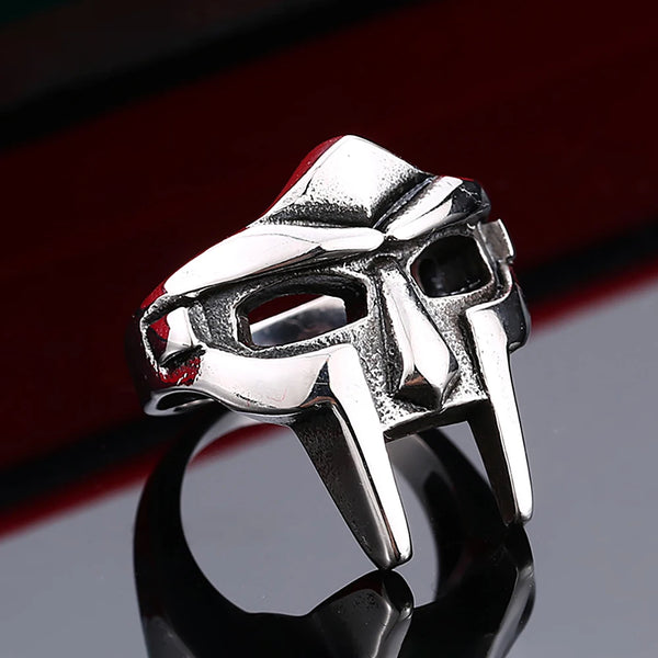 A silver stainless steel mask ring featuring a stylized skull design, displayed on a dark reflective surface.