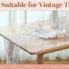 A wooden table is set with a white lace runner, glass vases, and pink flowers, suitable for vintage table decor.