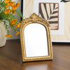 A vintage arched carved gold vanity mirror with a decorative frame sits on a wooden surface.