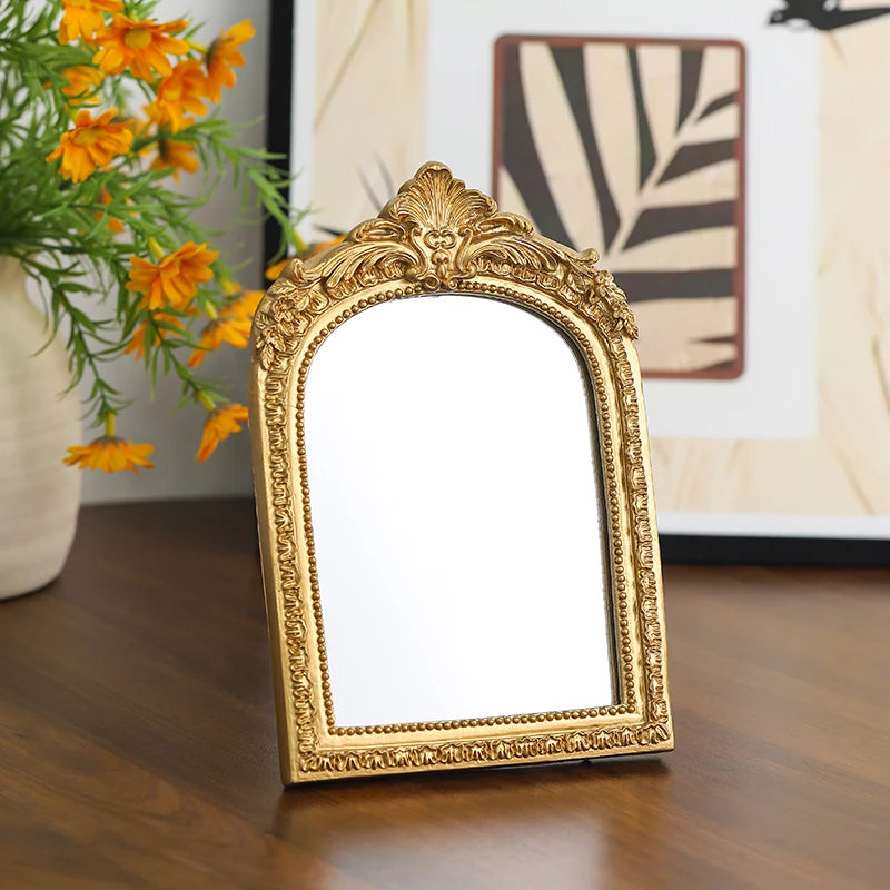 A vintage arched carved gold vanity mirror with a decorative frame sits on a wooden surface.