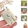 A hand holds a pressed metal flower, surrounded by dried flowers and tools for crafting.