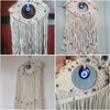 A macrame wall hanging featuring a white pendant with a blue evil eye and dangling threads.