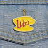 Yellow enamel pin shaped like a stylized 'L' with the word Luke's in red text pinned to denim fabric.