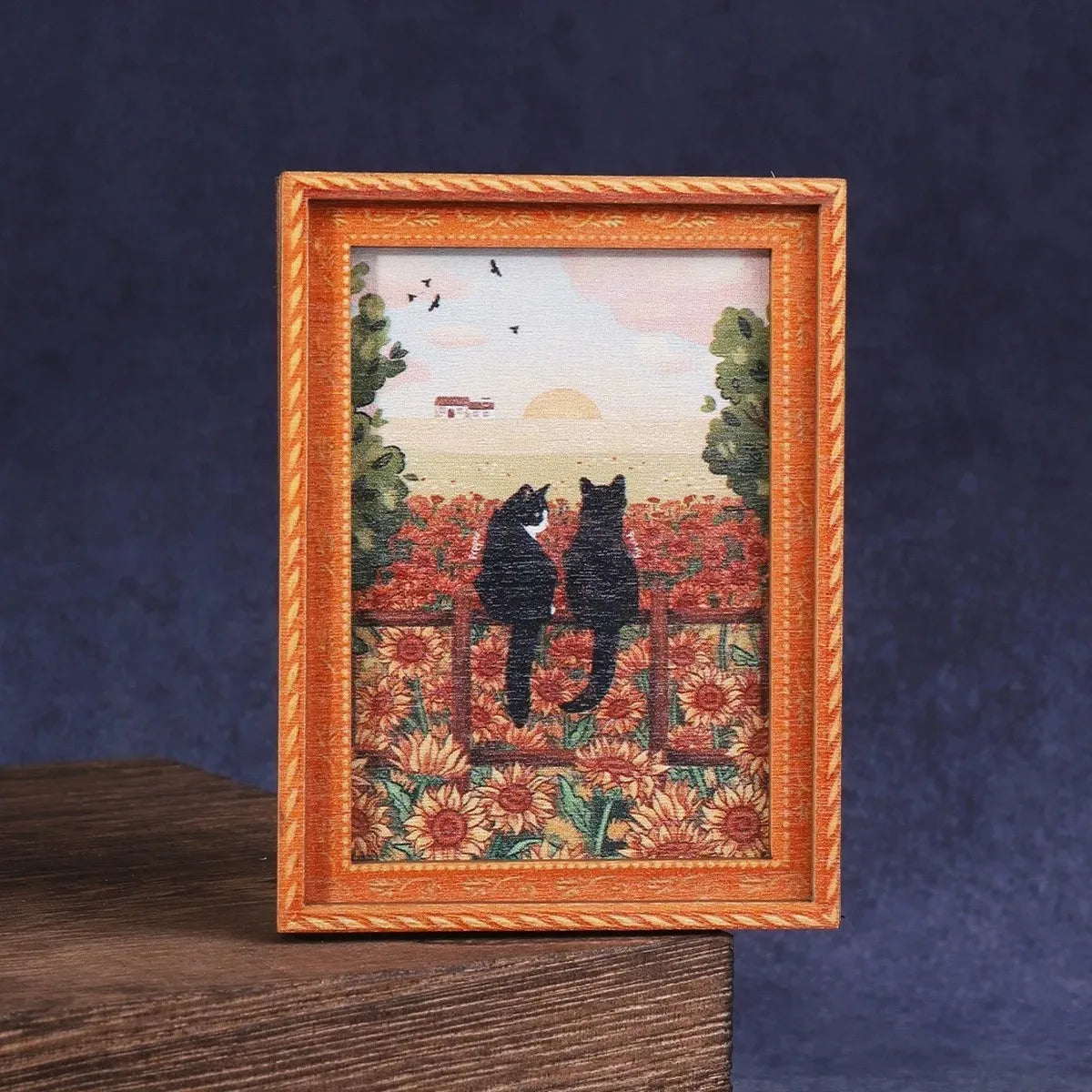 Una obra de arte enmarcada con dos gatos entre flores rojas y naranjas y un fondo de atardecer.