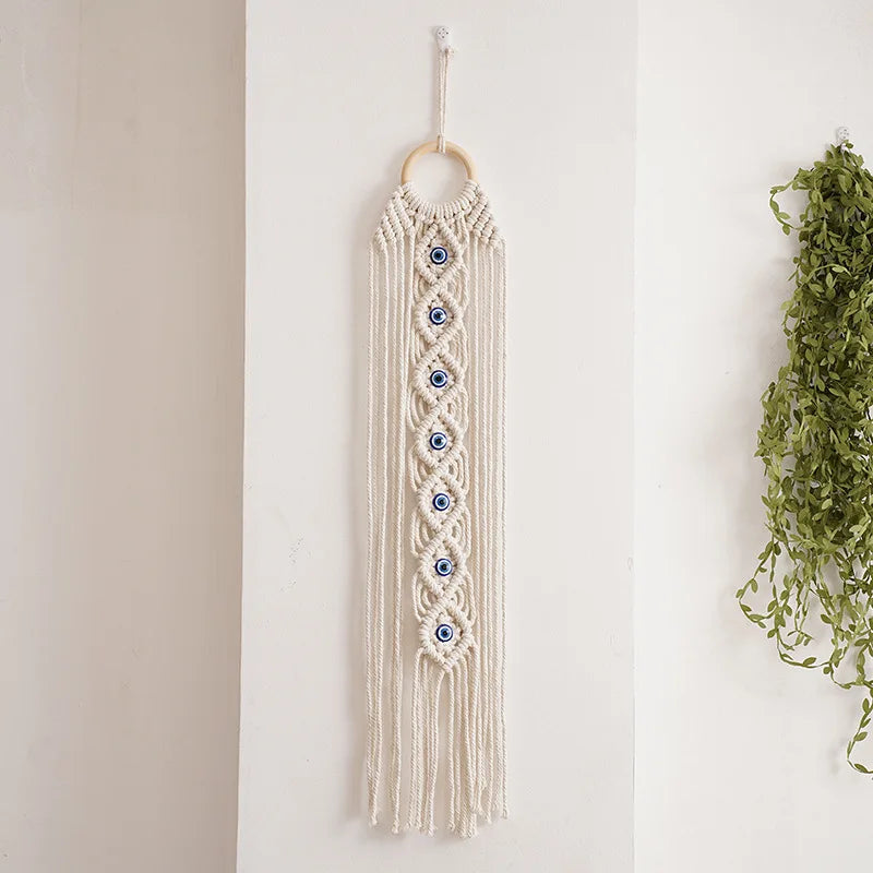 A cream-colored macrame wall hanging featuring a fringe and multiple blue evil eye pendants.