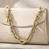 A light beige paperclip chain bag with a gold-toned chain strap is displayed on a surface.