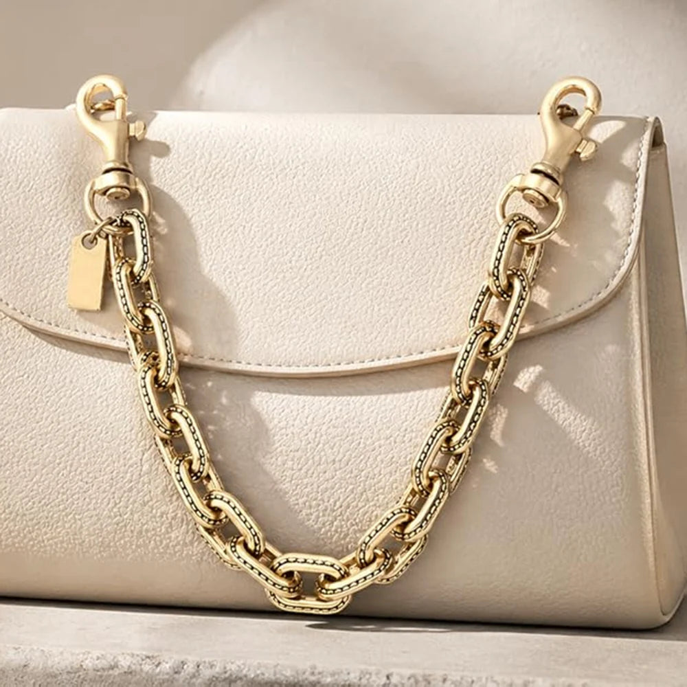 A light beige paperclip chain bag with a gold-toned chain strap is displayed on a surface.