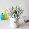 A white ceramic pot filled with silvery-white lavender artificial flowers sits on a white surface next to glass bottles.