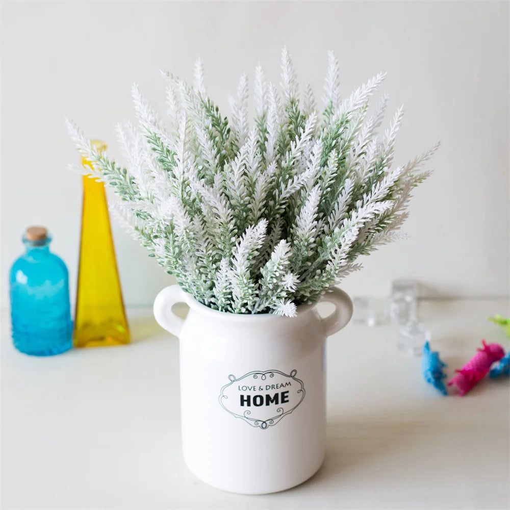 A white ceramic pot filled with silvery-white lavender artificial flowers sits on a white surface next to glass bottles.