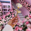 A hand holds an iridescent gold foil rose against a pink and white floral background.