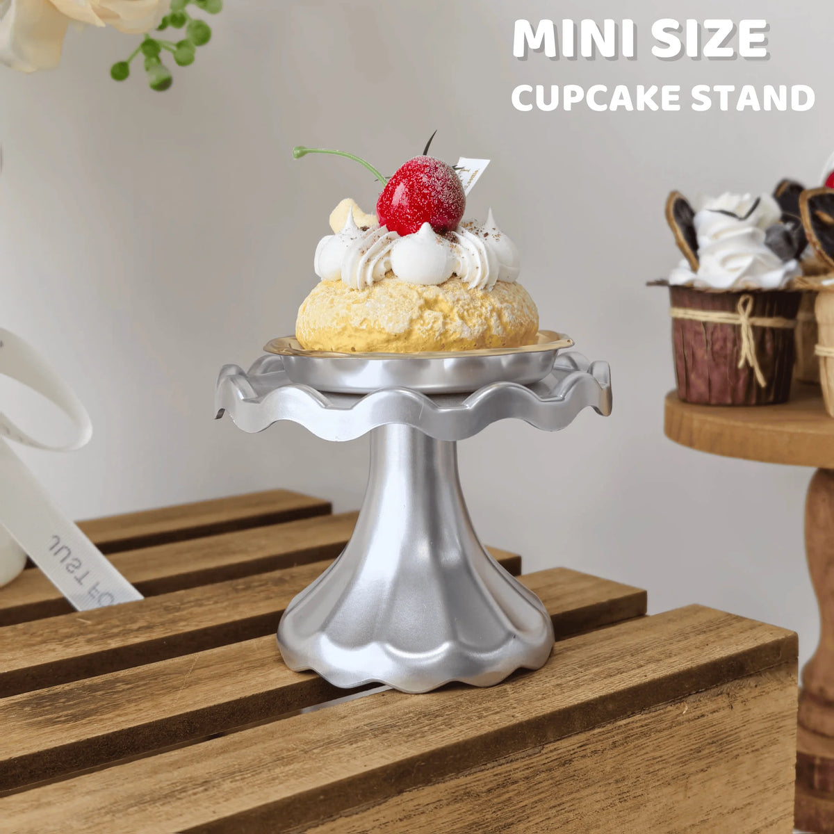 A mini silver cupcake stand displaying a single decorated cupcake with cream and berries on a wooden surface.
