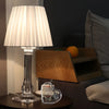A clear glass lamp with a pleated white shade sits on a dark table next to a wooden shelf and an acrylic heart-shaped decorat