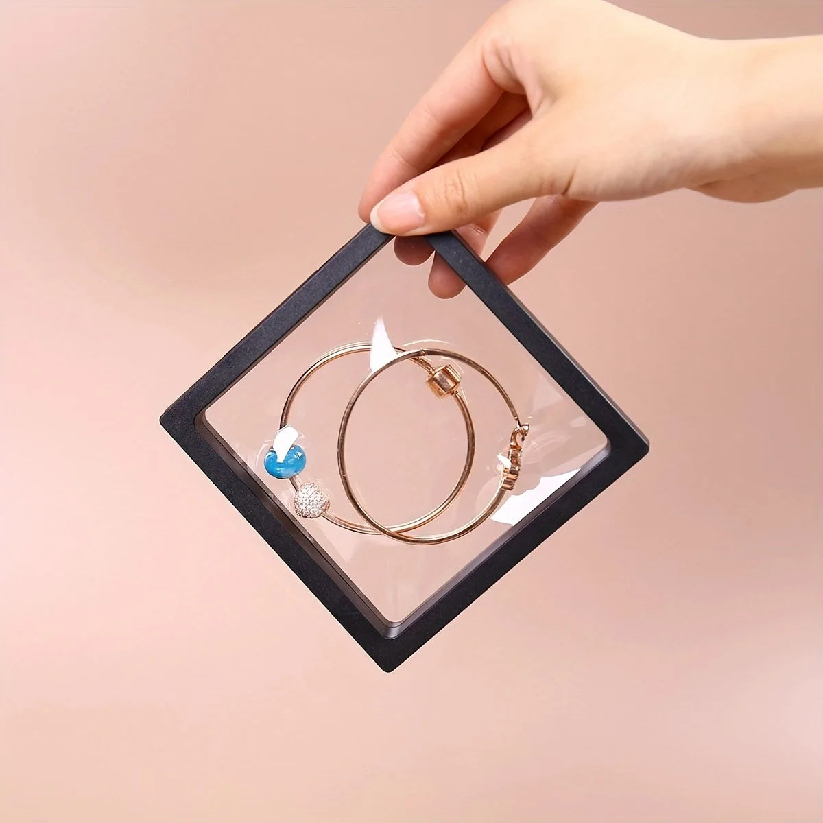 A hand holds a transparent display box with a geometric frame, showcasing gold and blue jewelry.