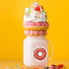 A pink milkshake glass filled with a creamy beverage, topped with whipped cream and colorful sprinkles, next to round donut l