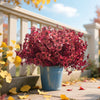 A vibrant pink and red artificial silk flower arrangement in a blue pot sits on a wooden deck with yellow autumn leaves.