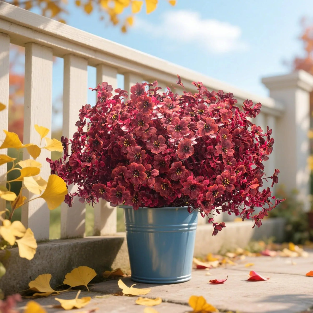 A vibrant pink and red artificial silk flower arrangement in a blue pot sits on a wooden deck with yellow autumn leaves.