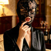 A woman wearing a black Venetian Gothic lace mask and a black satin garment looking directly at the camera.