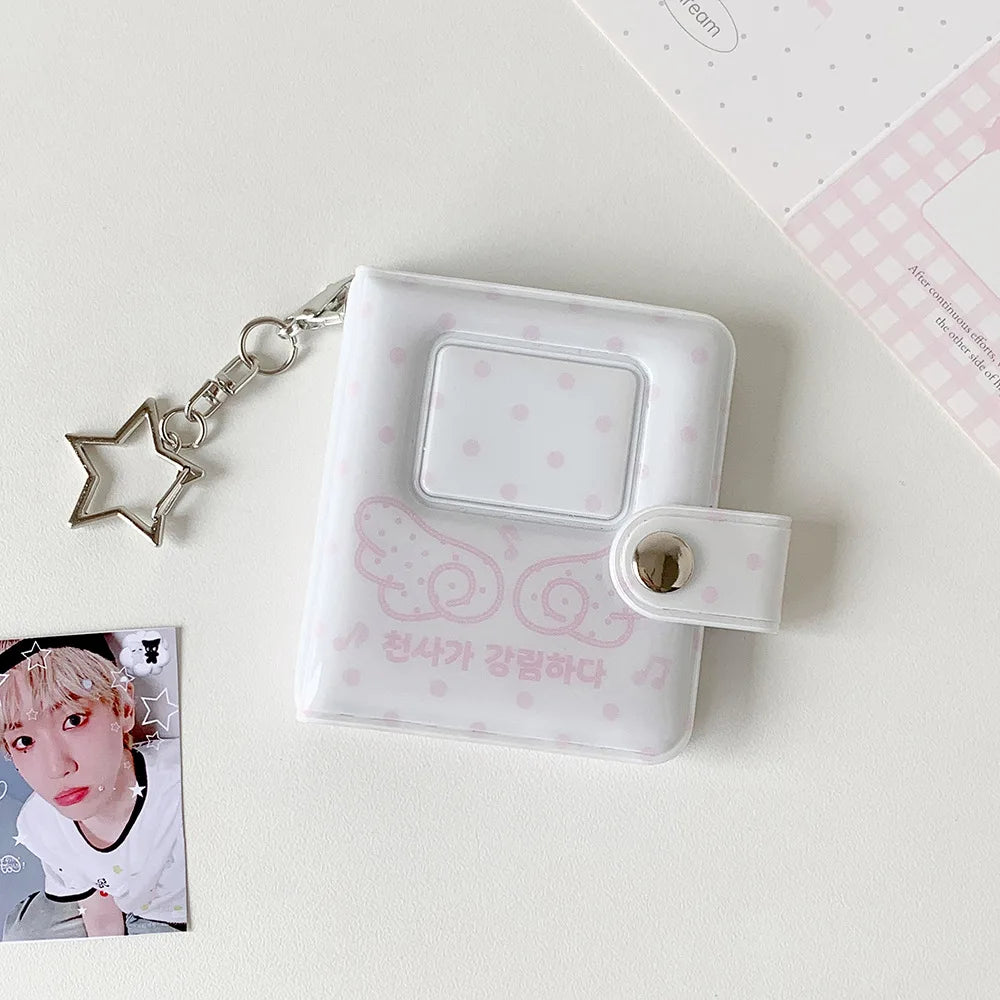 A white mini snap button photo album with a silver chain attached, resting on a light surface next to a K-pop card.