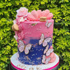 A vibrant pink and blue cake decorated with pink roses and gold butterfly toppers on a green foliage background.