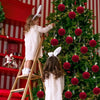 A young girl in a white dress is decorating a Christmas tree with red velvet flocked ornaments.