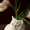 A white ceramic cat sculpture with long whiskers holds a bouquet of pink tulips.