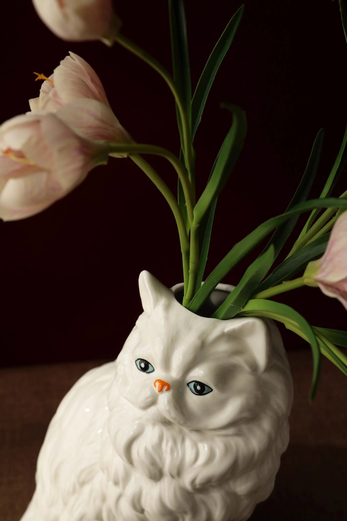 A white ceramic cat sculpture with long whiskers holds a bouquet of pink tulips.