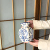 A hand holds a white porcelain vase with blue floral painting against a light paneled background.