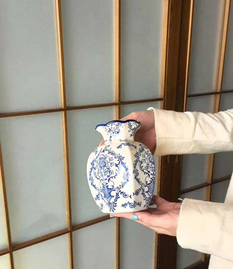 A hand holds a white porcelain vase with blue floral painting against a light paneled background.