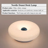 Eine runde, weiße, donutförmige Schreibtischlampe mit blauem Akzentlicht wird auf einer Oberfläche gezeigt.