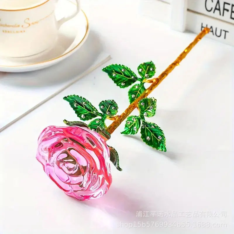 A crystal glass rose flower branch with green leaves resting on a white surface.