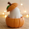A white elf figure is placed inside an orange pumpkin, decorated with a small green leaf and a brown stem.