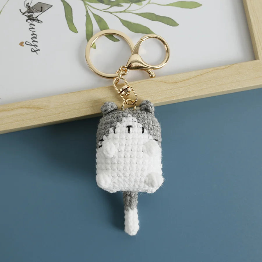 A handcrafted crochet cat keychain pendant with gray and white yarn, hanging from a gold-colored split ring.