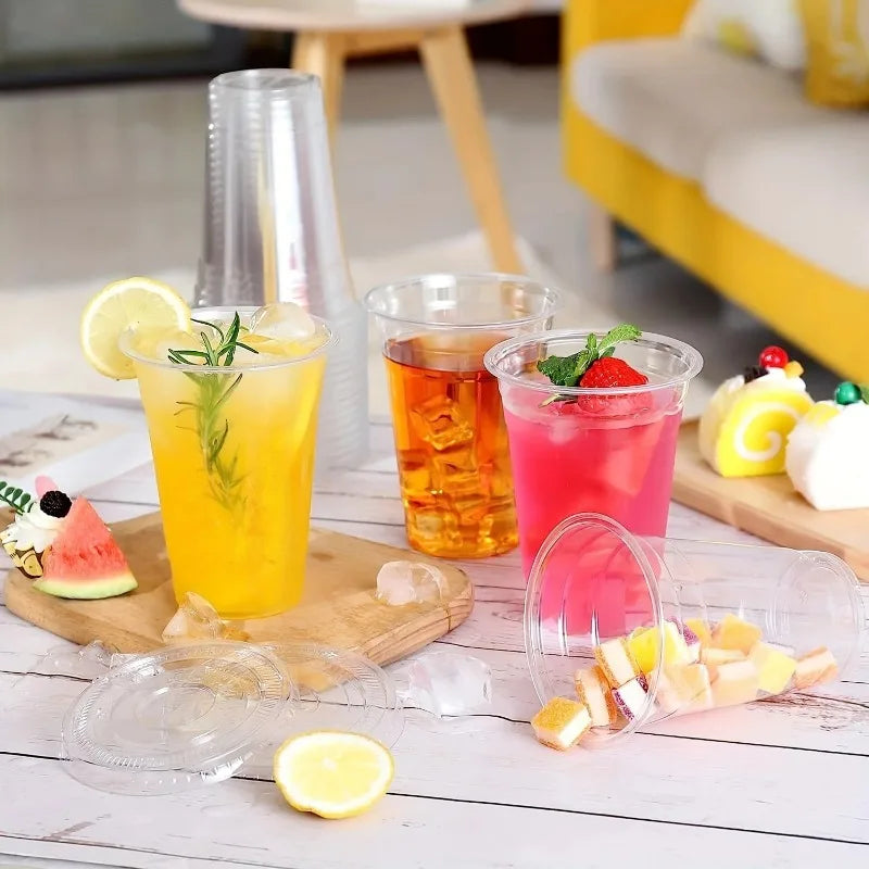 A collection of colorful citrus and fruit drinks in clear disposable plastic cups with flat lids on a white wooden surface.